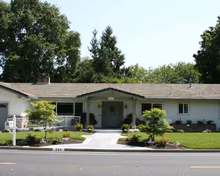 A single-story care home with a front entrance, landscaped lawn, and trees.