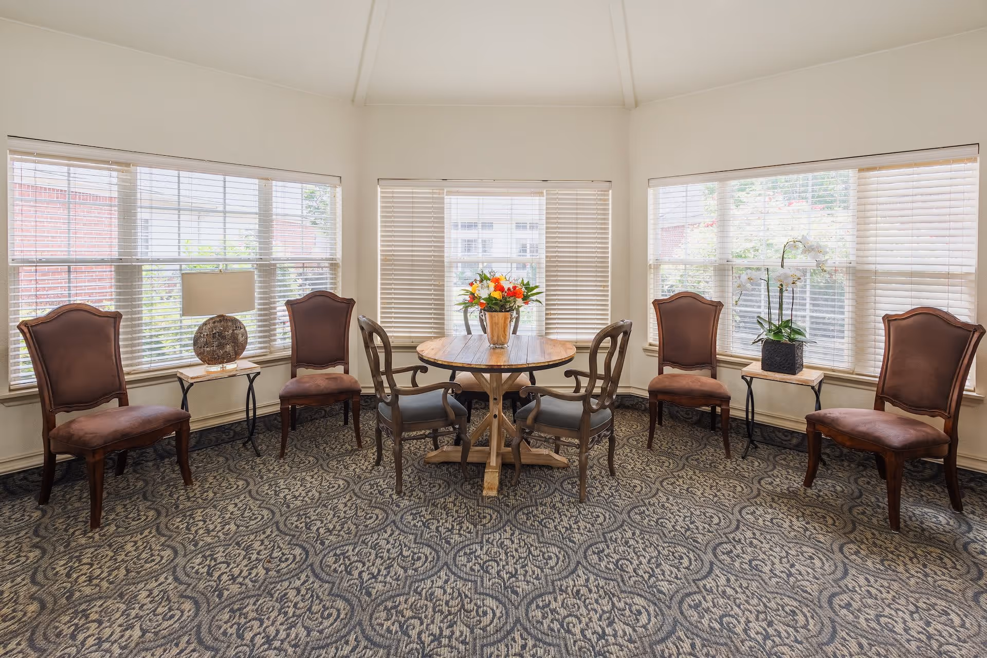 A bright room with large windows on three sides, featuring a round wooden table with a flower arrangement in the center. Surrounding the table are four wooden chairs with cushioned seats, and along the walls are four additional upholstered chairs. Two small side tables hold a lamp and a potted orchid. The floor is covered with a patterned carpet.