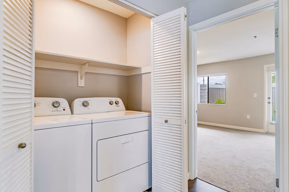 A laundry closet with side-by-side washer and dryer behind louvered bifold doors opening to a light, neutral living area.