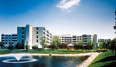 A large multi-story senior living facility building with white and light-colored exterior walls, surrounded by green lawns, trees, and a small pond with a fountain in the foreground under a blue sky with some clouds.