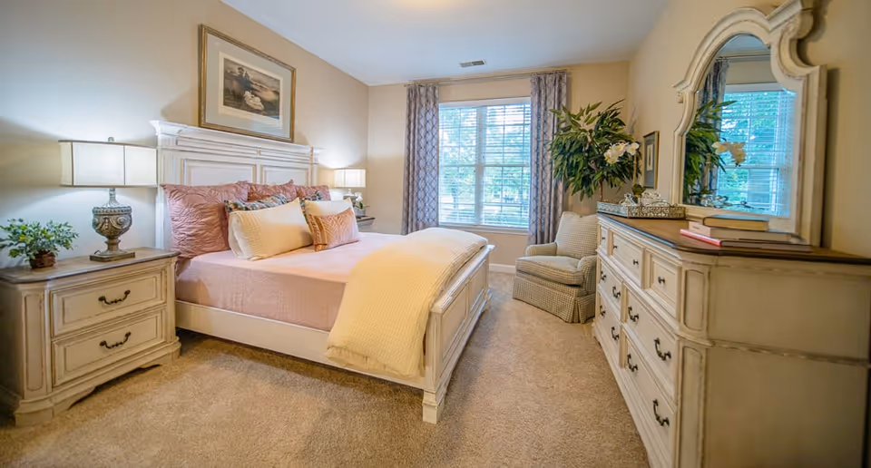 Bright traditional bedroom with a white bed, matching nightstands and dresser with mirror, an armchair, and a large window.