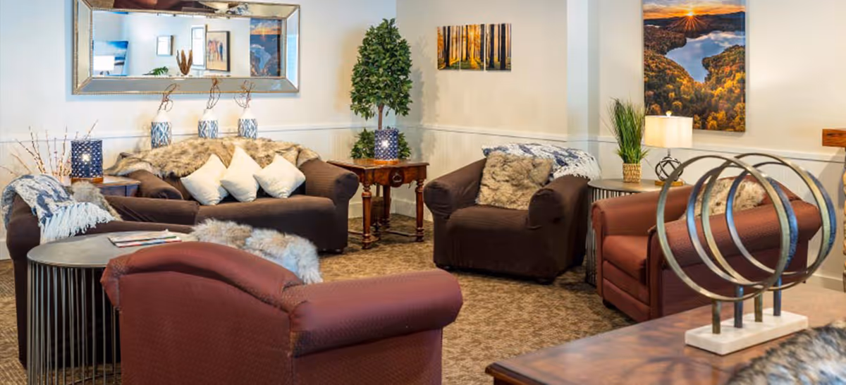 A cozy living room area with multiple armchairs and sofas in brown and maroon colors, decorated with white and fur throw pillows and blankets. The room features a large mirror on the wall, framed landscape artwork, a potted plant, side tables with lamps, and a decorative sculpture on a wooden table in the foreground.