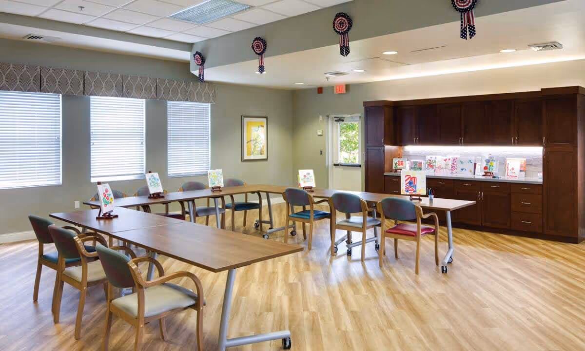 A bright and spacious activity room with wooden floors, several tables arranged in a U-shape, and chairs around them. The tables display colorful artwork on small easels. The room has large windows with blinds, a cabinet with additional artwork on the countertop, and patriotic decorations hanging from the ceiling.