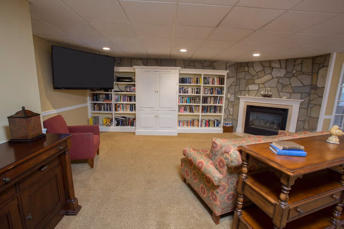 A cozy living room area featuring a stone fireplace, a large flat-screen TV mounted on the wall, built-in white bookshelves filled with books, a red armchair, a floral patterned armchair, and a wooden table with books and decorative items on it. The room has beige carpet and recessed lighting in the ceiling.