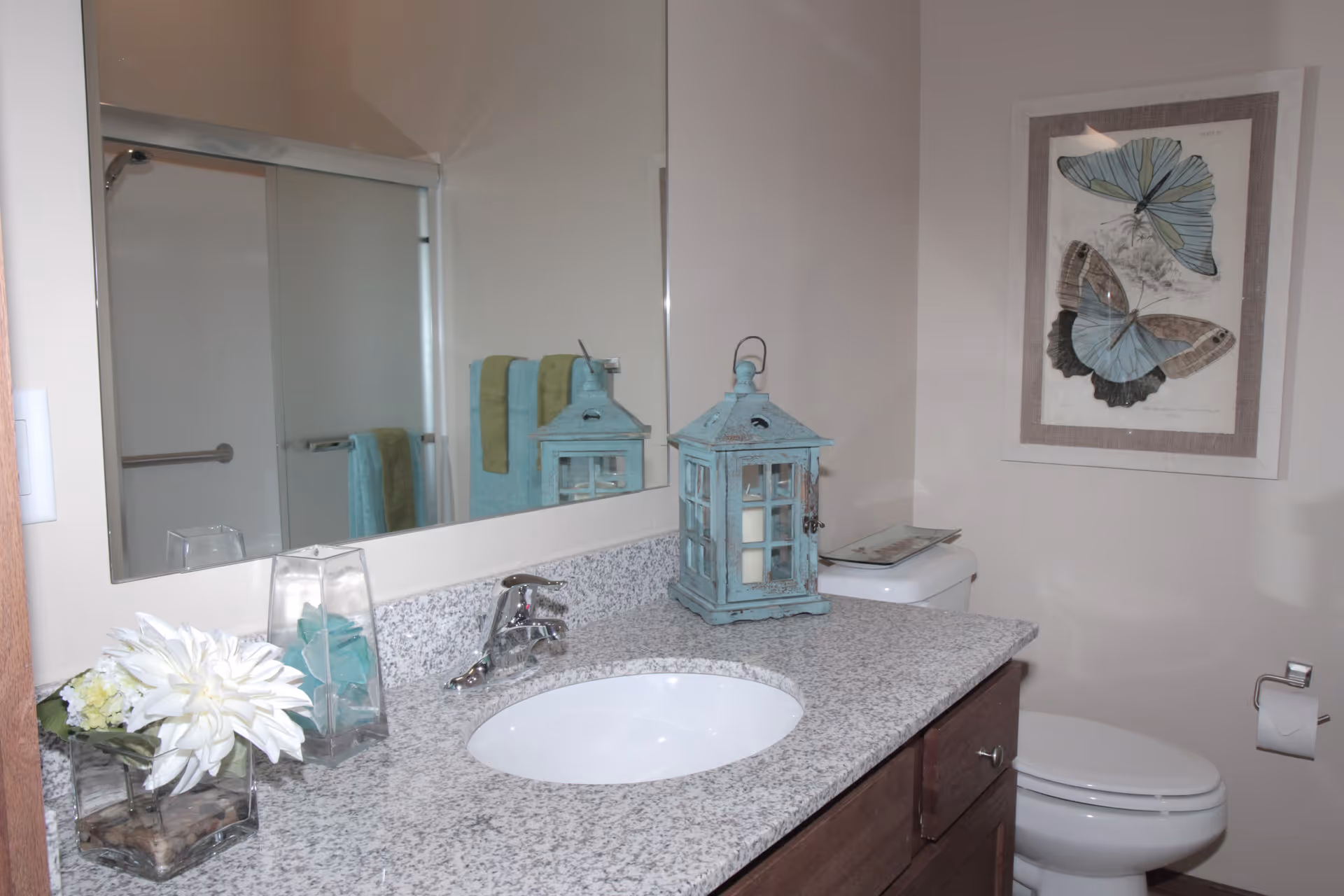 A bathroom with a granite countertop featuring a white sink and silver faucet. On the countertop, there is a decorative blue lantern, a glass vase with white flowers, and a clear container with blue items. A large mirror is mounted above the sink, reflecting a towel rack with blue and green towels. In the background, there is a white toilet with a framed butterfly artwork hanging on the wall above it. A toilet paper holder with a roll of toilet paper is mounted on the wall beside the toilet.