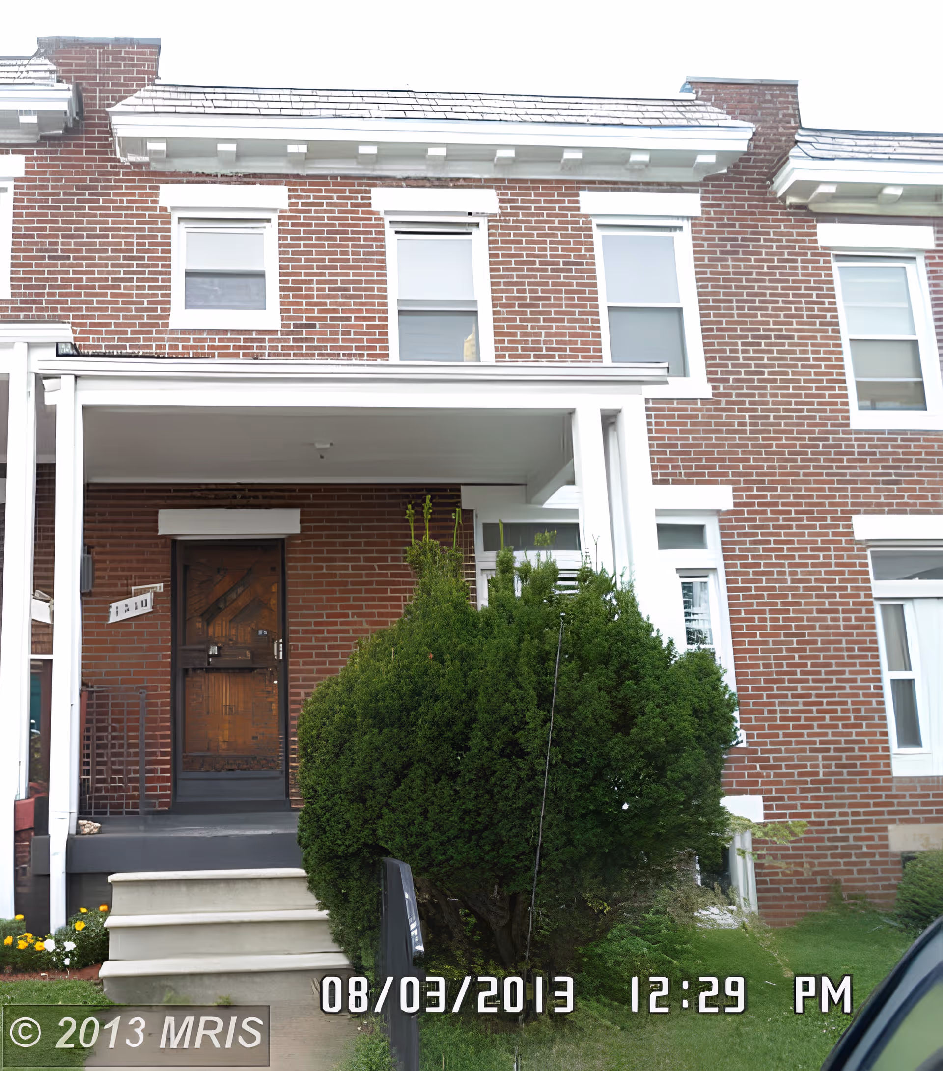 Front exterior view of a two-story brick townhouse with white trim. There is a small covered porch with steps leading up to a wooden front door. A large green bush is in front of the porch, and the lawn is neatly maintained.
