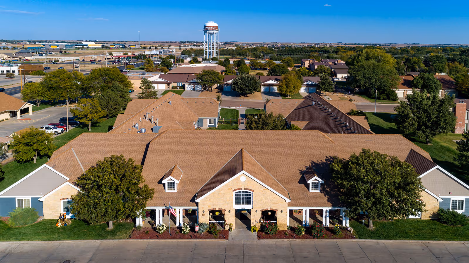 Aerial front view of a single-story brick assisted living facility with a central entrance, landscaped beds, and surrounding parking and neighborhood.