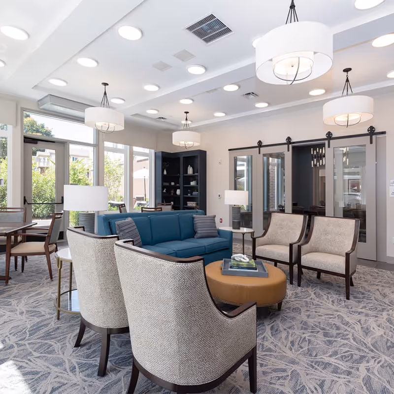 A bright and modern senior living community lounge area with a blue sofa, four beige armchairs, a round brown ottoman with a tray on top, and several tables with lamps. Large windows let in natural light and offer a view of greenery outside. The ceiling features multiple round recessed lights and hanging drum pendant lights. There is a dark bookshelf in the corner and sliding glass doors with dark frames on one wall.