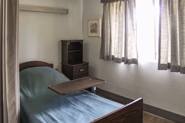 Small single-occupancy bedroom with a hospital-style bed, overbed table, nightstand, and a curtained window.