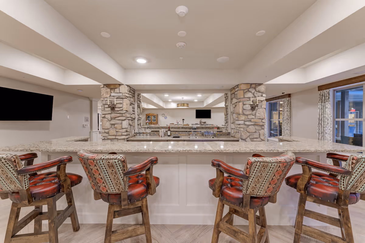 Interior view of a senior living facility's bar area with a granite countertop and four wooden bar stools upholstered with patterned fabric and red leather seats. The space features stone pillars, a large mirror behind the bar, two flat-screen TVs mounted on the walls, and windows with patterned curtains allowing natural light to enter.