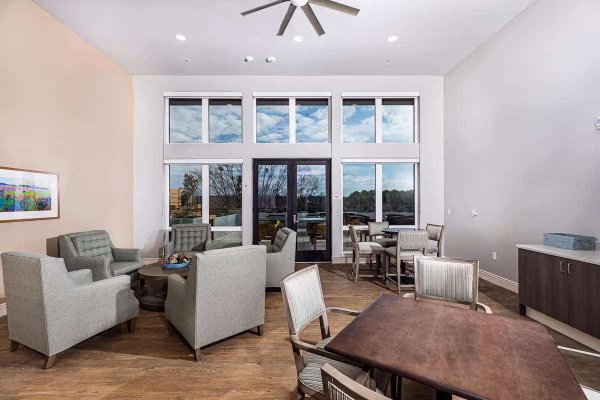 A bright and spacious common area with large windows and glass doors letting in natural light. The room features a seating area with four gray upholstered armchairs around a round wooden coffee table, and two dining tables with chairs. The floor is wooden, and there is a ceiling fan above. A colorful framed artwork is hung on the left wall.