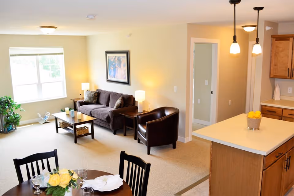 A bright and cozy living area in a senior living facility featuring a brown sofa with cushions, a dark brown leather armchair, a coffee table with candles, and two side tables with lamps. There is a large window with blinds letting in natural light, a framed picture on the wall, and a small dining table with chairs and a floral centerpiece. The kitchen area with wooden cabinets and a countertop with a basket of lemons is partially visible.