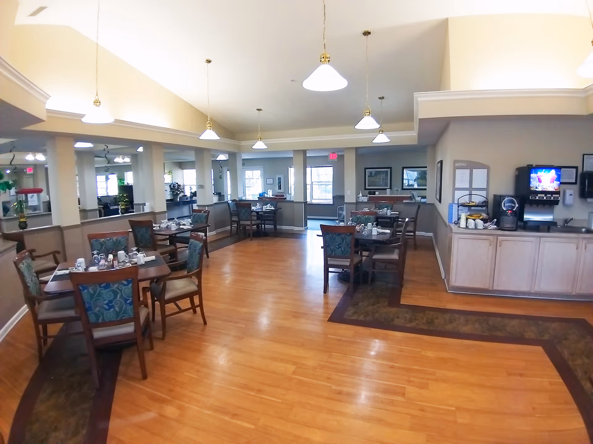 A dining room in a senior living facility with several wooden tables and chairs arranged neatly. Each table is set with cups, glasses, and napkins. The room has hardwood floors, beige walls, and multiple hanging pendant lights. On the right side, there is a counter with a coffee machine, a beverage dispenser, and a menu board on the wall. Large windows allow natural light to enter the space.