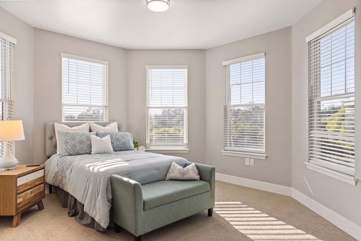 A bright and airy bedroom with a large bed adorned with multiple pillows and a light blue comforter. There is a small green upholstered bench at the foot of the bed with a decorative pillow. The room has beige walls, carpeted floor, and four large windows with white blinds letting in natural light. A wooden nightstand with a lamp is positioned next to the bed.