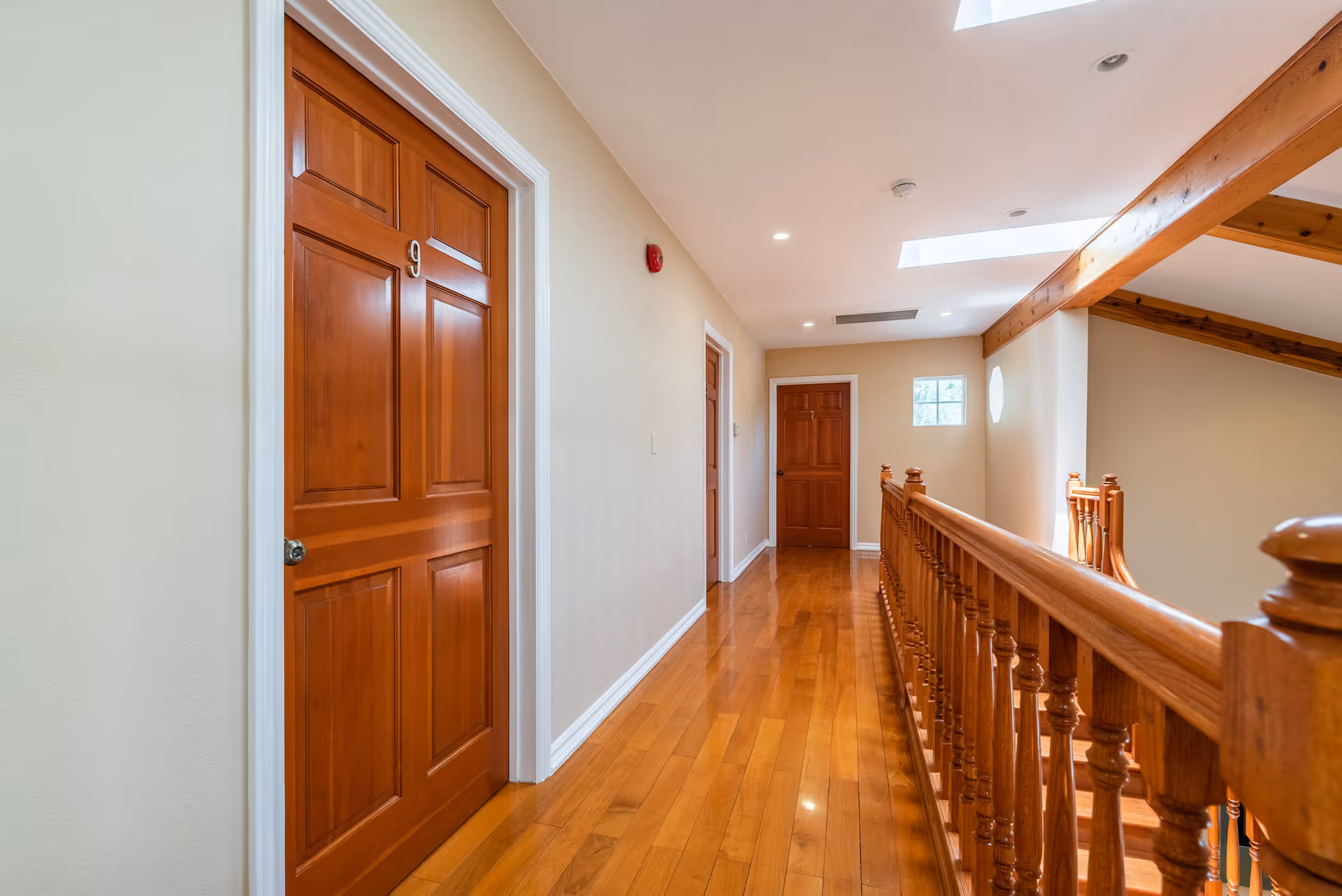 A well-lit indoor hallway with polished wooden floors and wooden doors, one door labeled with the number 9. The hallway has beige walls, recessed ceiling lights, a wooden railing on the right side, and a small window at the end letting in natural light.