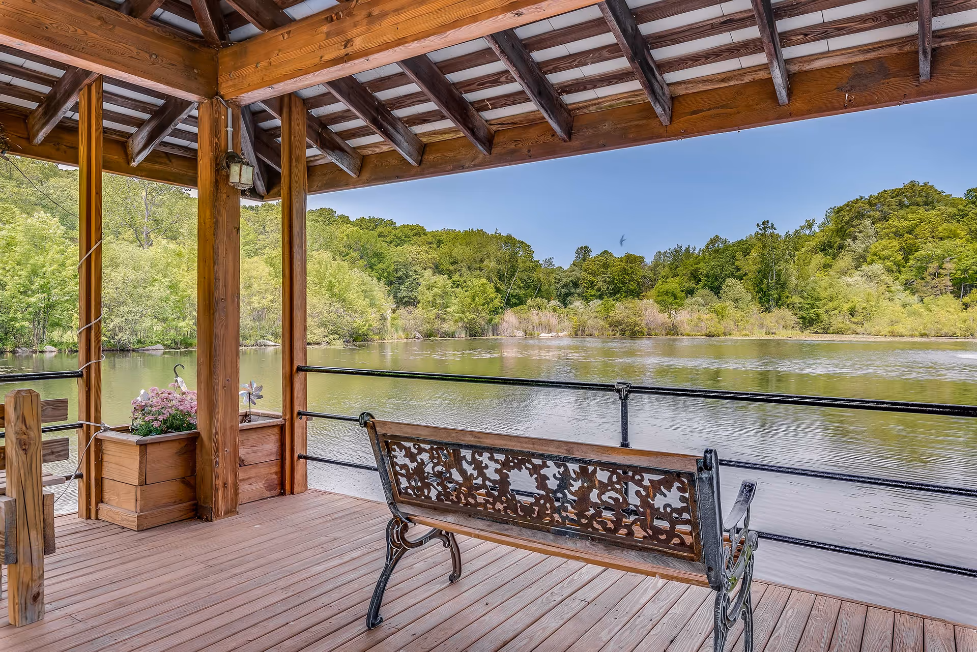 A wooden covered deck overlooking a calm lake surrounded by green trees under a clear blue sky. The deck has a decorative metal and wood bench facing the water and a wooden planter with pink flowers.