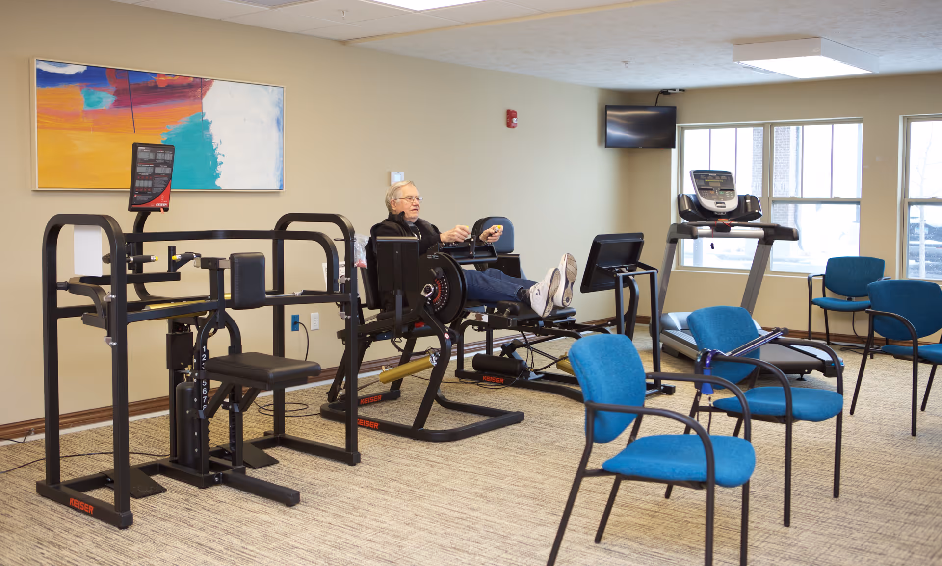 An elderly man exercising on a seated rowing machine in a fitness room with various exercise equipment, blue chairs, a wall-mounted TV, and large windows letting in natural light. A colorful abstract painting hangs on the wall.