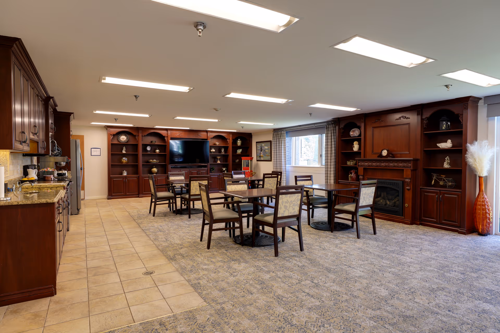 A senior living facility common area with a kitchen on the left featuring wooden cabinets and granite countertops. Several tables with chairs are arranged on a carpeted floor in front of a large wooden entertainment center with a flat-screen TV and decorative items. There is a fireplace integrated into the wooden shelving unit on the right side, and a window with curtains allowing natural light into the room.