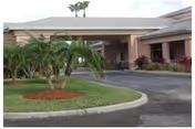 Exterior view of the entrance to Atlantic Shores Nursing and Rehab Center showing a covered driveway with a circular landscaped area featuring palm trees and other greenery.