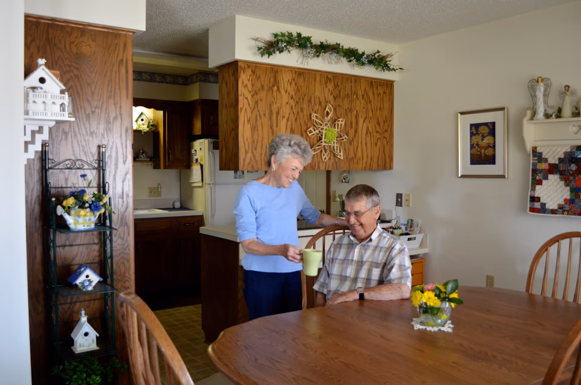 An elderly woman hands a mug to an elderly man seated at a wooden dining table in a homey kitchen-dining area.