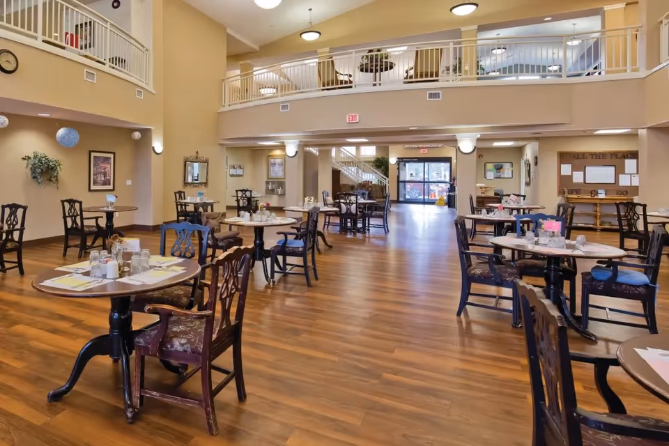 Spacious senior living dining room with multiple round tables and chairs arranged on a wooden floor. The room features high ceilings with a second-floor balcony railing, warm beige walls, and large windows near the entrance allowing natural light. Decorative plants, framed pictures, and a clock adorn the walls.