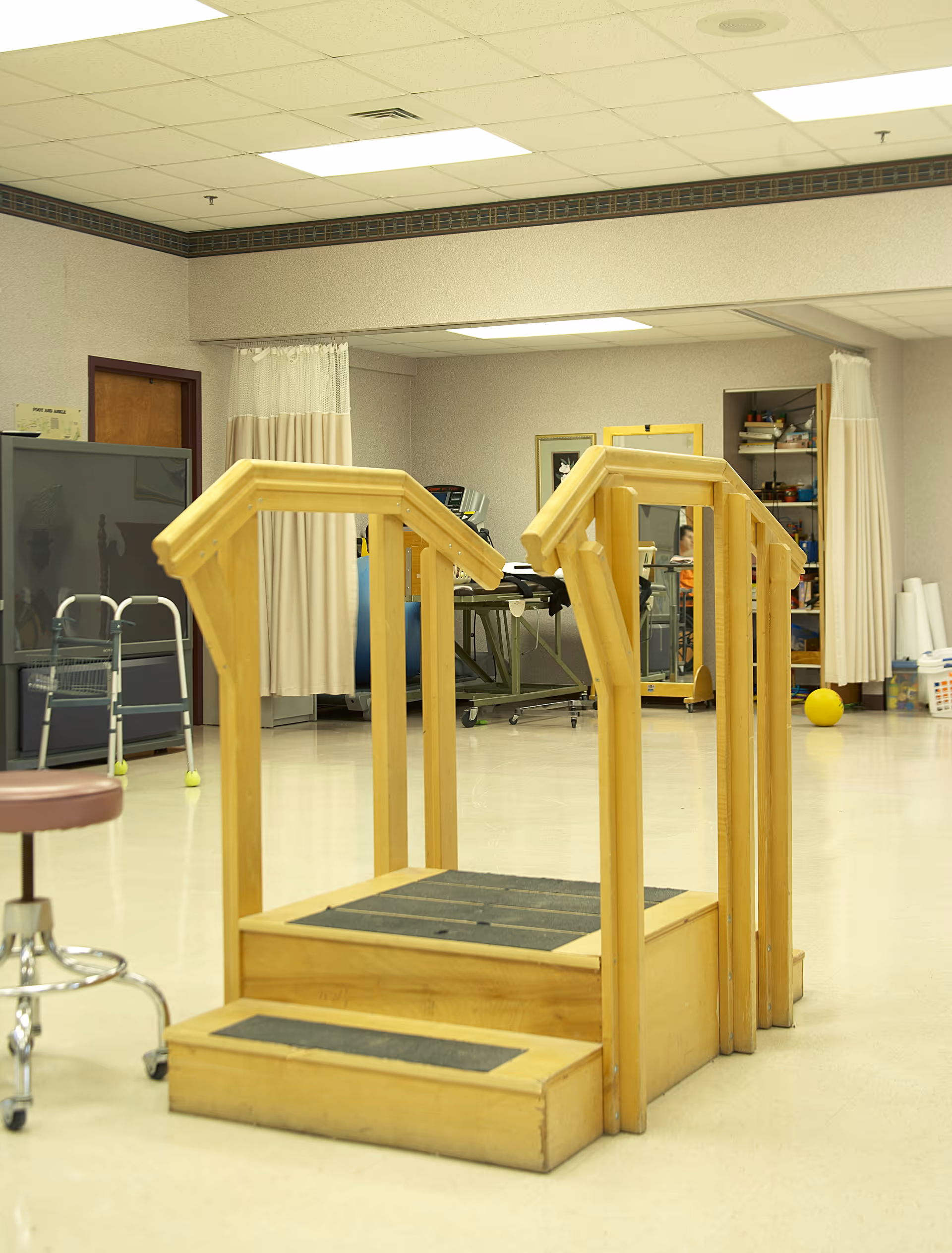 Indoor physical therapy room featuring wooden practice stairs with handrails and exercise equipment in the background.