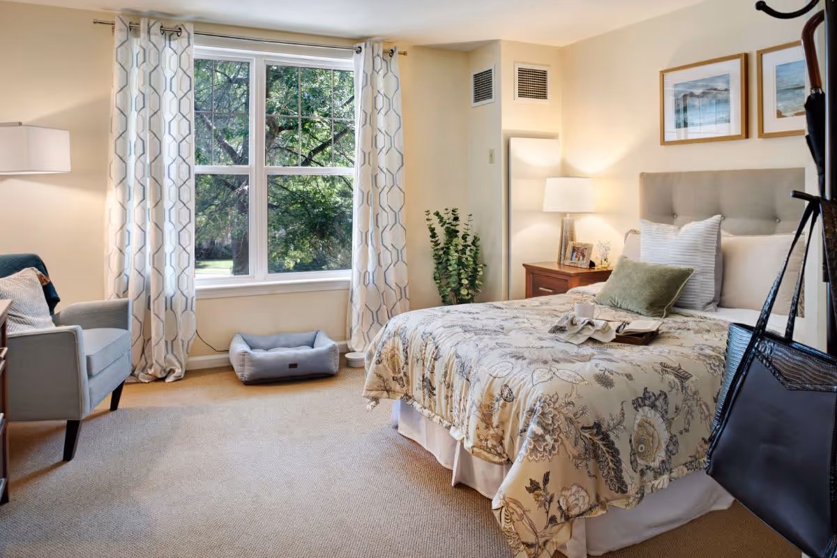 A cozy bedroom with a large window showing green trees outside. The room features a bed with floral bedding and multiple pillows, a wooden nightstand with a lamp and framed photos, a gray armchair with a pillow, patterned curtains, a pet bed on the floor near the window, and a coat rack with a black bag hanging on it.