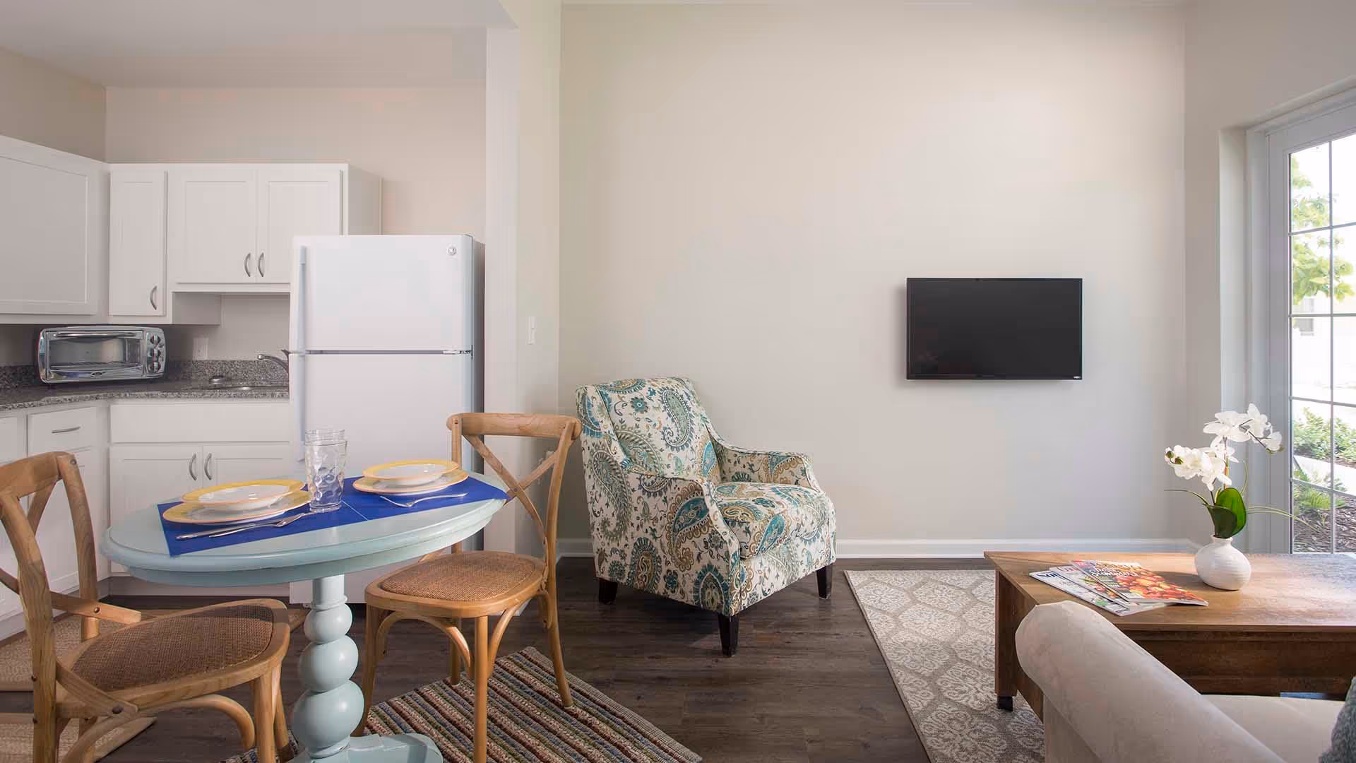 A cozy senior living community interior featuring a small dining area with a round table set for two, wooden chairs, a white kitchen with cabinets, a refrigerator, and a toaster oven. Adjacent to the kitchen is a patterned armchair, a wall-mounted flat-screen TV, a wooden coffee table with magazines and a white vase with flowers, and a large window letting in natural light.