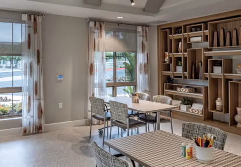 A bright and modern interior room with large windows covered by sheer curtains, two tables with chairs around them, and a wooden shelving unit filled with decorative items and plants. The tables have art supplies like colored pencils and glue sticks on them.