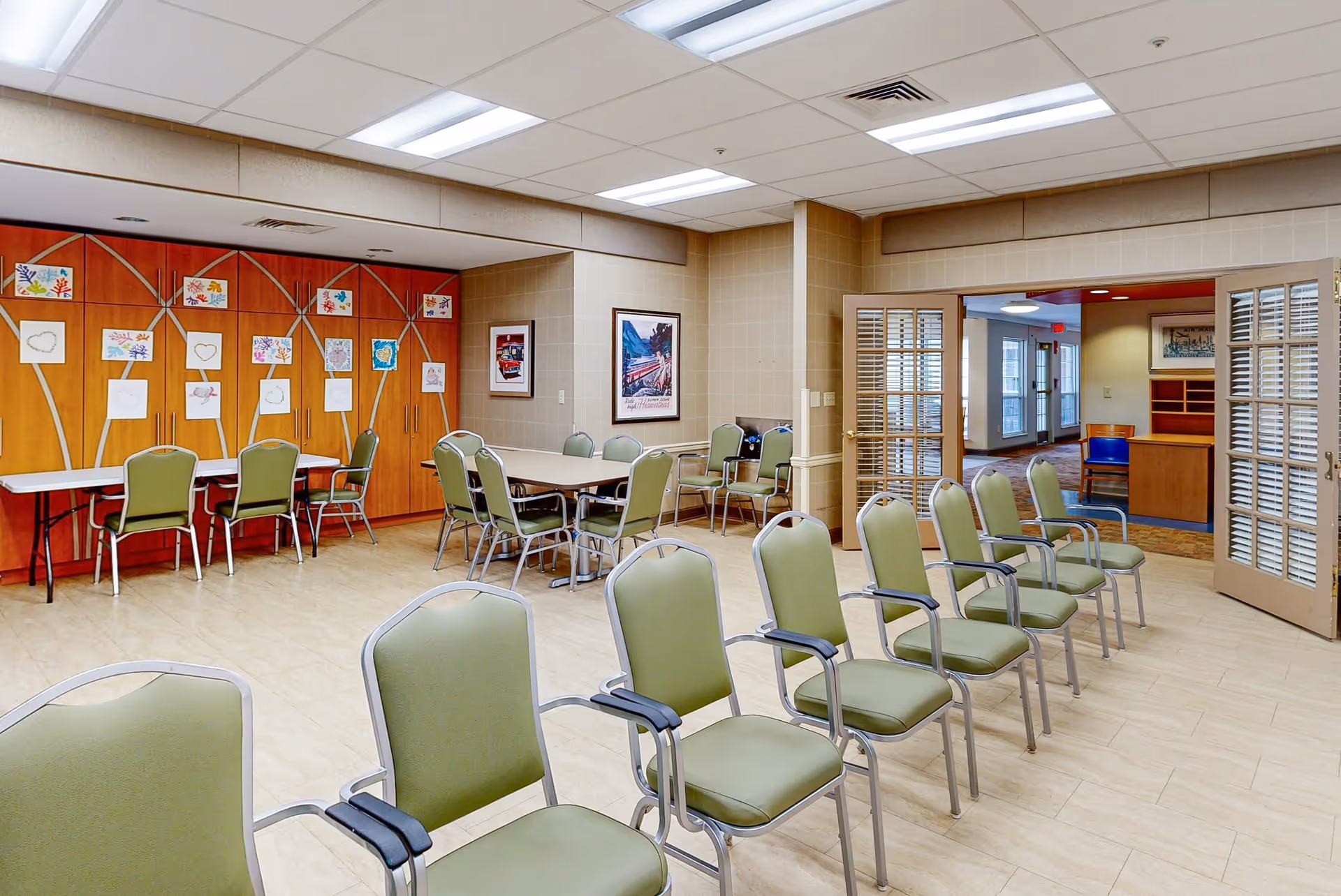 A spacious room with rows of green chairs arranged in two lines facing forward. There are tables and additional chairs along the back wall, which is decorated with children's artwork and drawings. The room has beige tiled walls and a light-colored floor, with double doors leading to another area of the facility.