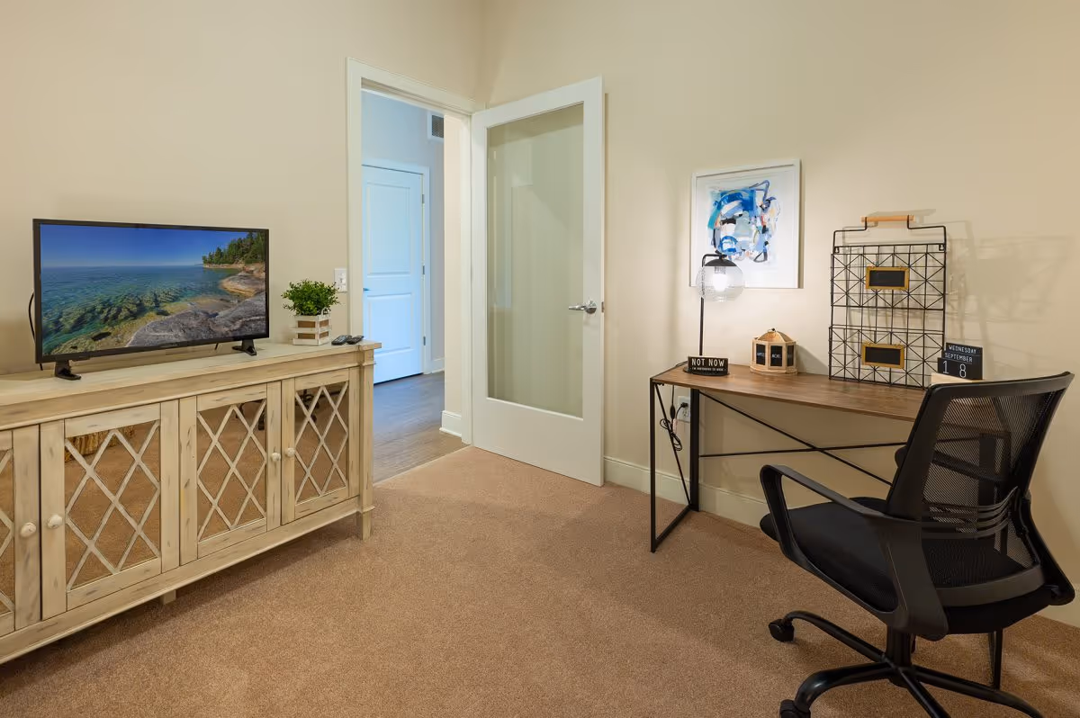 A small room with beige walls and carpeted floor featuring a wooden cabinet with a flat-screen TV displaying a nature scene, a small potted plant, and a remote control on top. There is a glass-paneled door open to an adjacent room with wooden flooring. On the right side, there is a wooden desk with a black mesh office chair, a desk lamp, decorative items, and a framed abstract artwork on the wall.