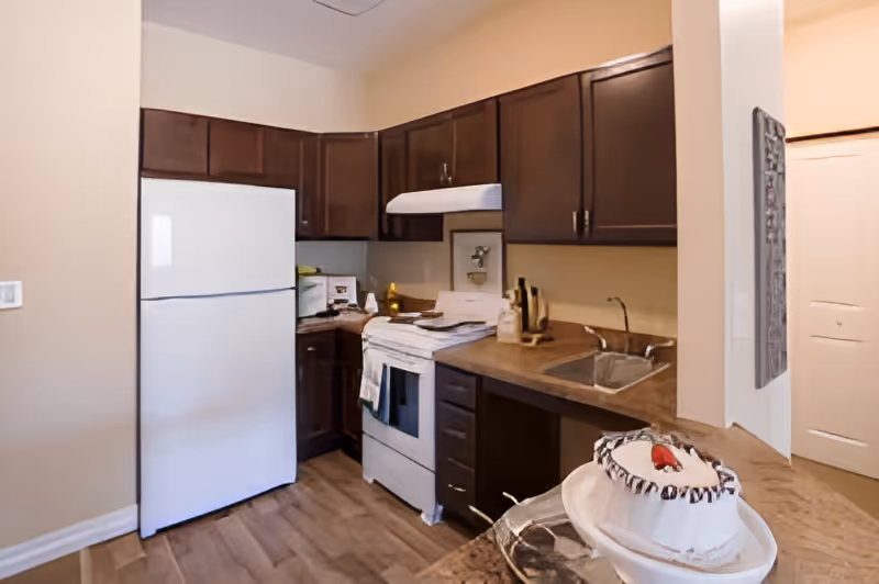 A kitchen area in a senior living facility featuring dark brown cabinets, a white refrigerator, a white stove with a range hood, a sink, and a countertop with a cake on a plate in the foreground.