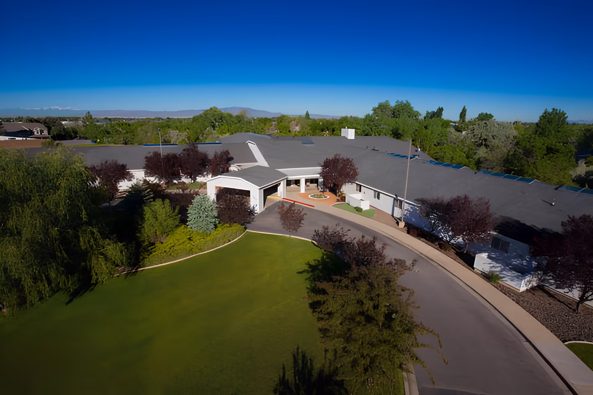 Aerial view of Apple Tree Assisted Living facility showing a large single-story building with a gray roof surrounded by green trees and landscaped lawns under a clear blue sky.