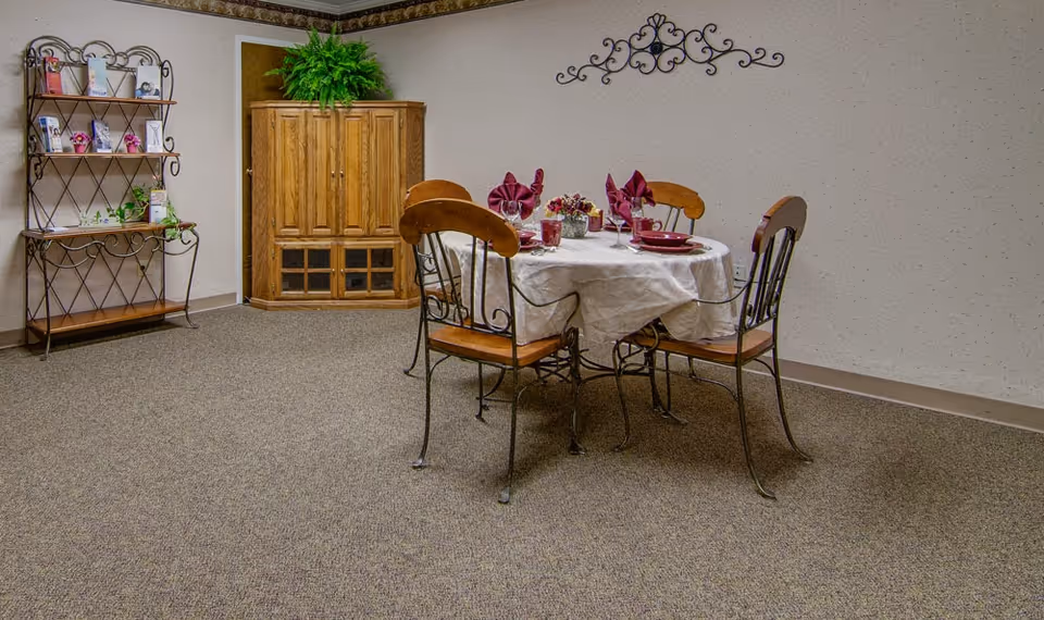 A small dining area with a round table covered with a white tablecloth, set with red napkins, plates, and glasses. Four metal chairs with wooden seats surround the table. In the background, there is a wooden cabinet with a green plant on top and a metal shelf unit with decorative items and plants. The walls are light-colored with a decorative metal wall hanging above the table.