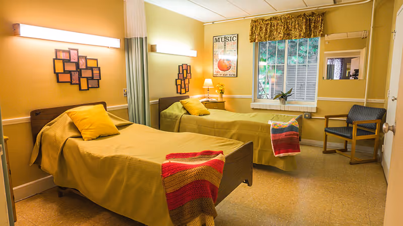 A cozy room with two single beds, each covered with a yellow bedspread and a colorful knitted blanket at the foot. Each bed has a yellow pillow. The room has warm yellow walls with framed artwork, a window with a floral valance, a wooden chair, and a small bedside table with a lamp and a vase of flowers.