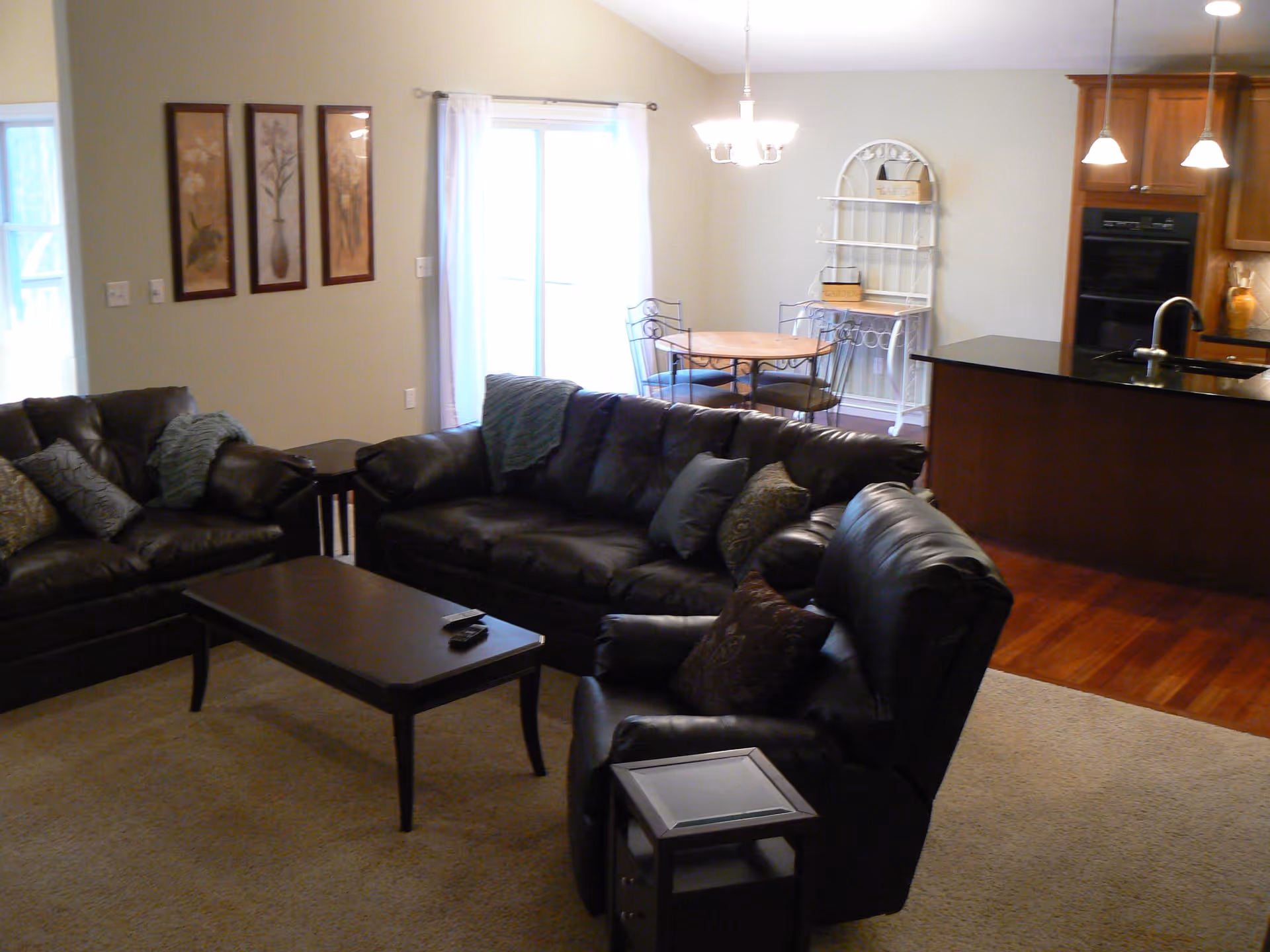 A cozy living room with dark leather sofas and armchairs arranged around a dark wooden coffee table. Behind the living area is a small dining space with a round table and four chairs, next to a window with white curtains. To the right, there is a kitchen area with wooden cabinets, a black countertop, and pendant lights hanging from the ceiling.
