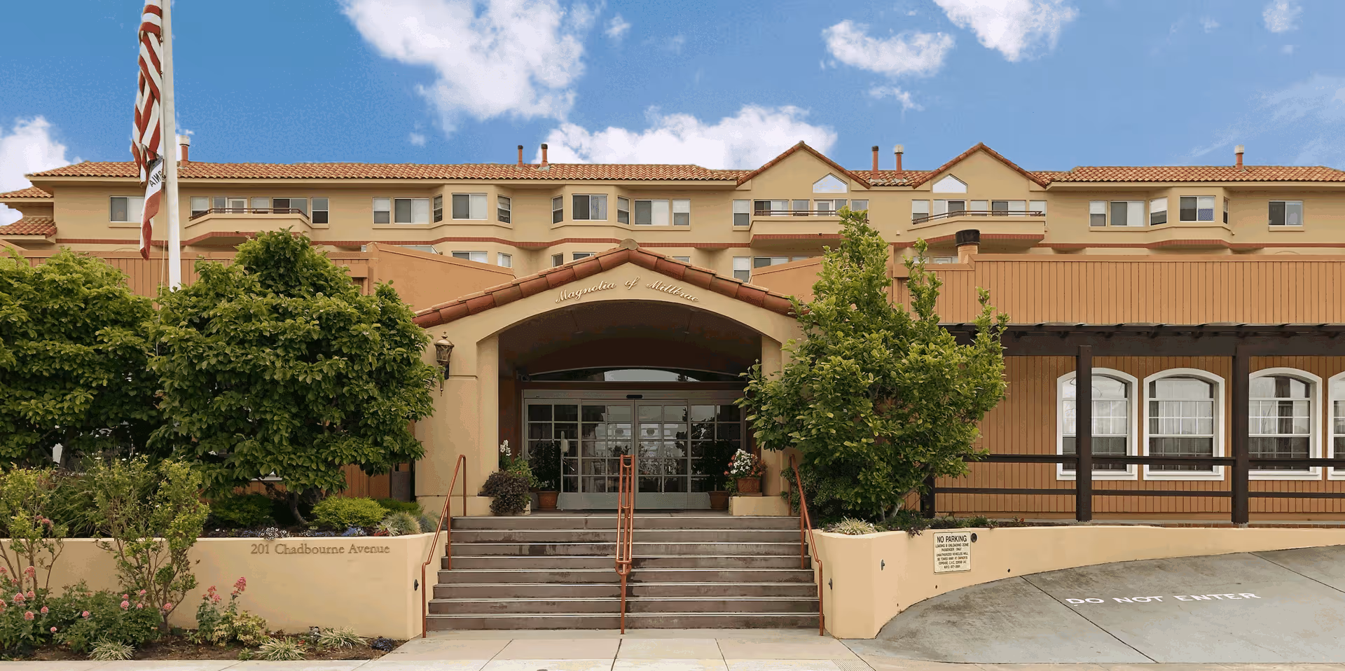 Front entrance of The Magnolia of Millbrae senior living facility with steps, landscaping, and an American flag.