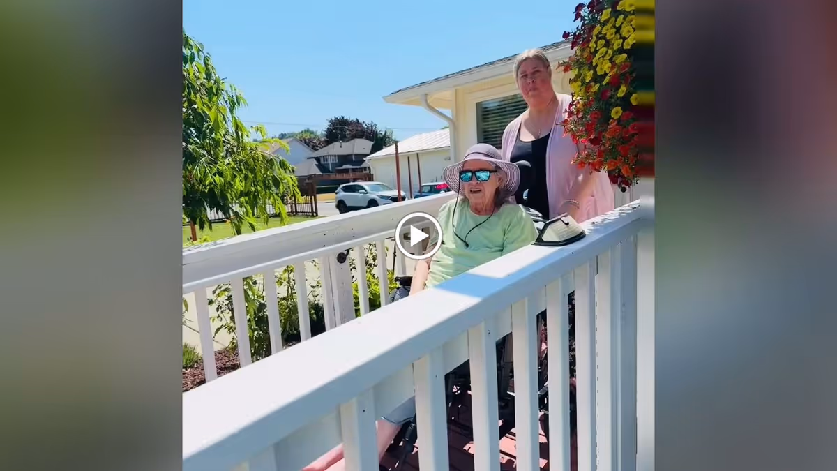 An elderly person wearing a sunhat and sunglasses sits in a wheelchair on a white wooden ramp outside a building. A caregiver stands behind them. There are colorful hanging flowers on the right side and a tree and parked cars in the background under a clear blue sky.