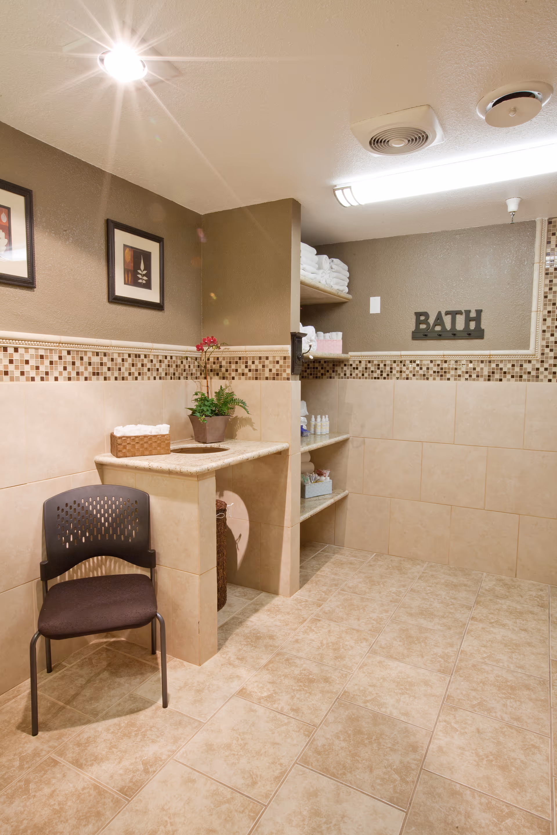 A clean and well-lit bathroom area with beige tiled walls and floor. There is a black chair with a brown cushion in front of a small counter holding a basket with towels and a potted plant. Built-in shelves on the right side hold neatly folded white towels and various toiletries. The word 'BATH' is displayed on the wall above the shelves. Two framed pictures hang on the left wall.