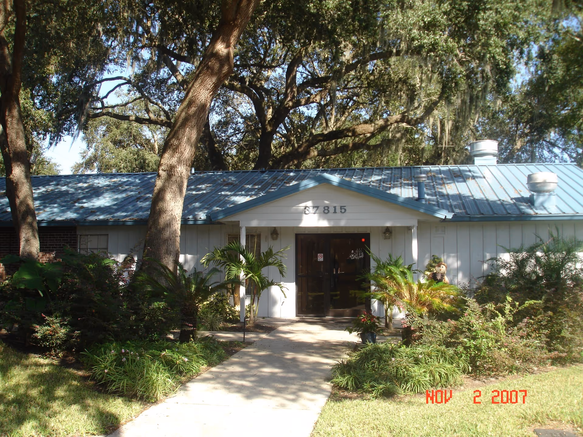 Front exterior view of a single-story building with a blue metal roof and white siding, surrounded by trees and greenery. A concrete pathway leads to the entrance with glass double doors. The building number 37815 is displayed above the entrance. The date stamp on the image reads Nov 2 2007.