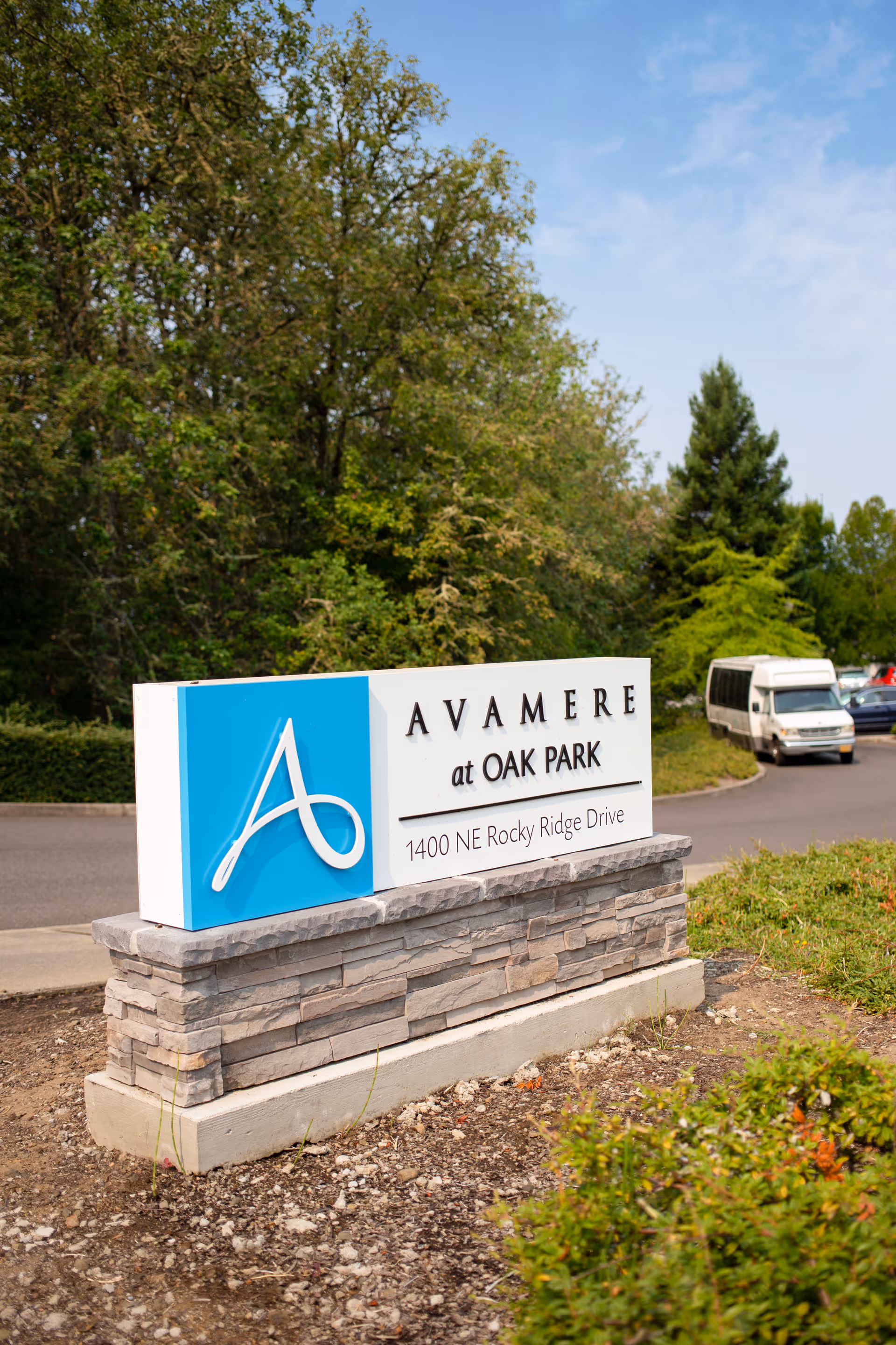 Outdoor stone sign for Avamere at Oak Park located at 1400 NE Rocky Ridge Drive, with trees and a road with parked vehicles in the background.