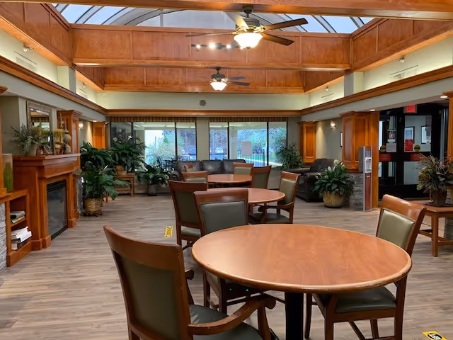 A spacious common area with round wooden tables and chairs, large windows letting in natural light, ceiling fans, wooden beams on the ceiling, a fireplace on the left side, and several potted plants around the room.