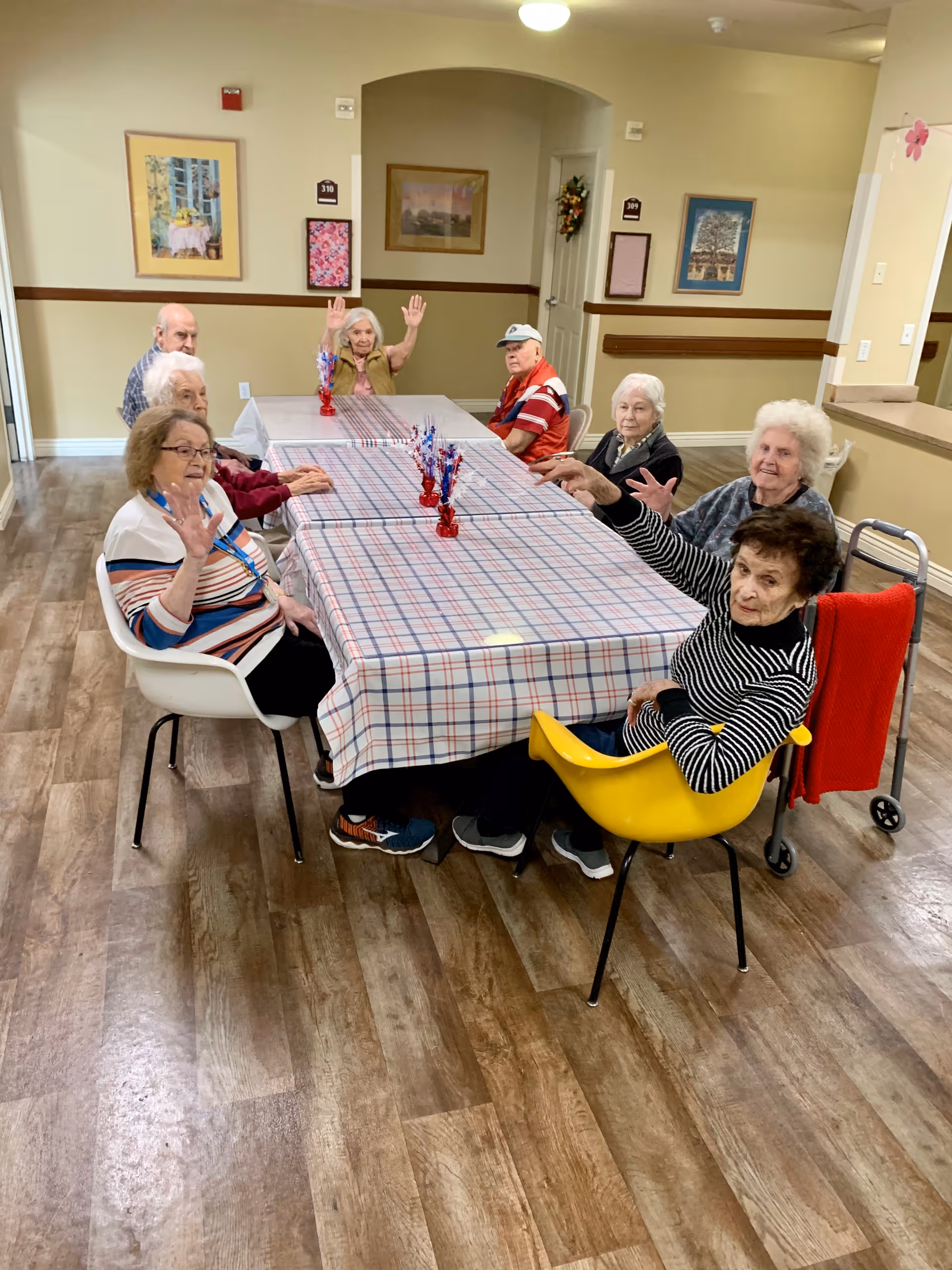 A group of elderly people sitting around a long table covered with a plaid tablecloth in a communal room. Some of them are waving at the camera. The room has wood flooring, framed pictures on the walls, and a doorway in the background.