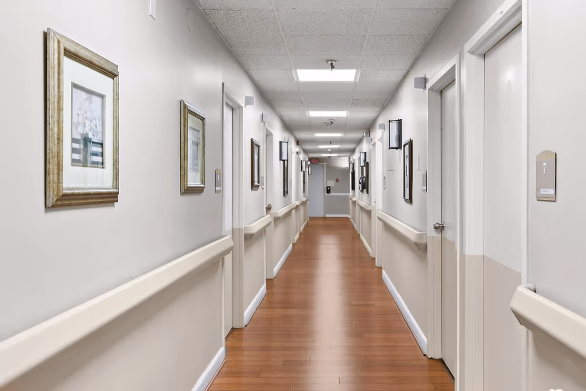 A long, clean hallway in a senior living facility with wooden flooring, beige walls, handrails on both sides, framed pictures hanging on the walls, and several closed white doors with room numbers.