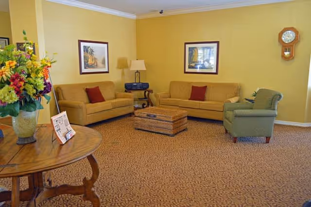 A cozy living room with two mustard yellow sofas, a green armchair, a wicker coffee table, and a wooden side table with a lamp and stereo. The walls are painted yellow and decorated with framed artwork and a wall clock. A round wooden table with a vase of colorful flowers is in the foreground.