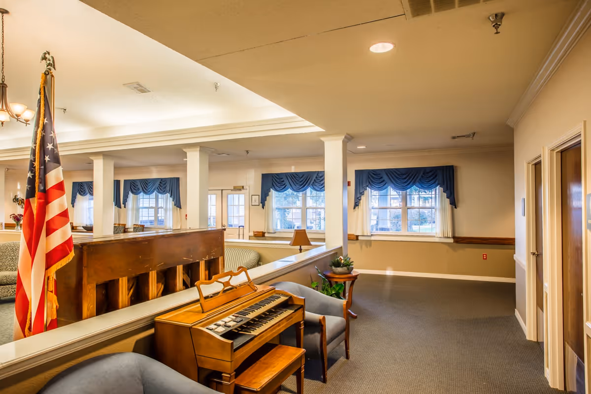 Community lounge with an organ, seating, an American flag, and windows dressed with blue valances.