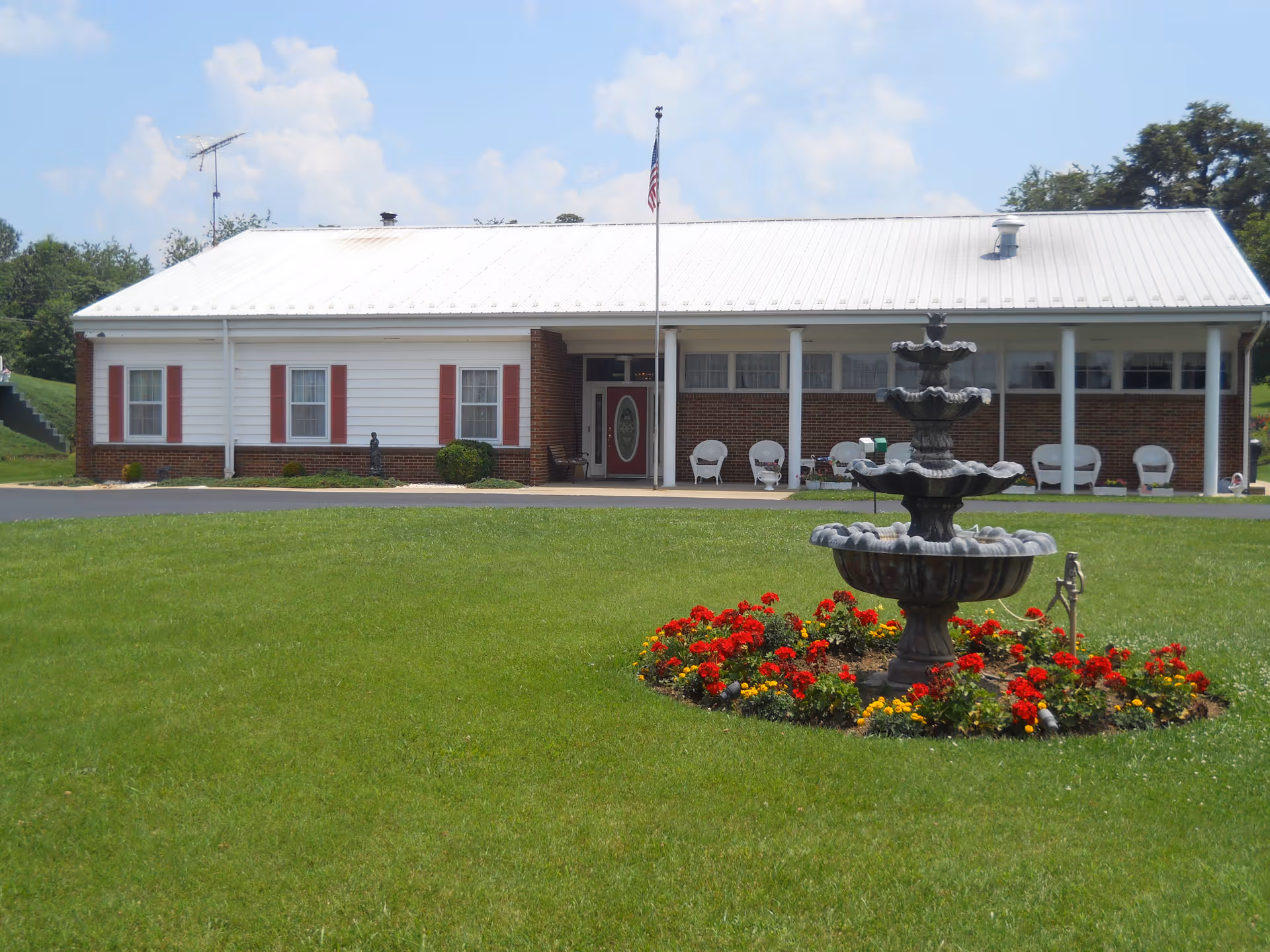 Single-story building with a white metal roof and brick lower walls, featuring a front porch with white columns and several white chairs. In front of the building is a green lawn with a tiered stone fountain surrounded by red and yellow flowers. An American flag is mounted on a pole near the entrance.