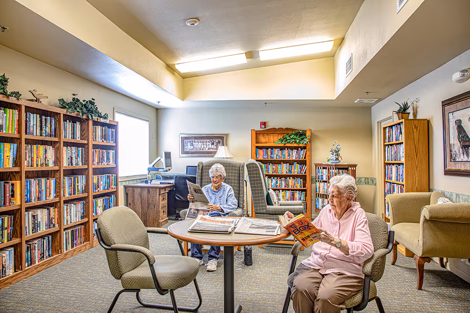 Two elderly women reading books and newspapers in a well-lit communal library/reading room with bookshelves and comfortable chairs.