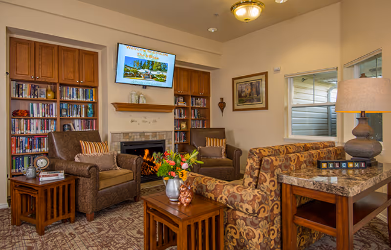 Comfortable senior living lounge with armchairs, patterned sofa, fireplace, bookshelves and a TV above the mantel.