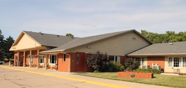 Exterior view of a single-story brick and siding building with a sloped roof, benches along the front porch, and a small landscaped area with bushes and a brick planter. The building is part of Accura HealthCare of Le Mars.